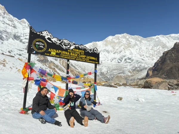 Three trekkers sitting under the welcome sign at Annapurna Base Camp with colorful prayer flags and snow-covered Annapurna massif in the background