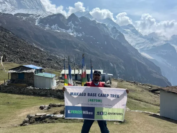 At Makalu Base Camp, a trekker is standing on a grassy field holding a “Makalu Base Camp Trek (4870m)” banner, with Mount Makalu visible in the background.