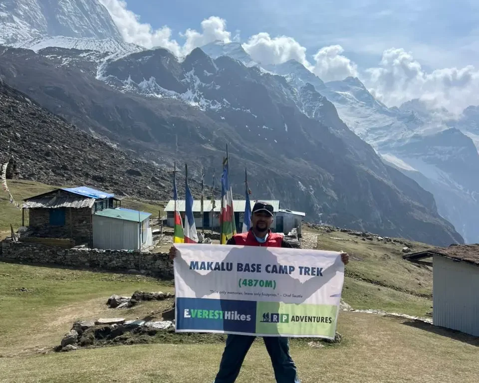 At Makalu Base Camp, a trekker is standing on a grassy field holding a “Makalu Base Camp Trek (4870m)” banner, with Mount Makalu visible in the background.