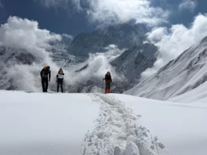 Three hikers trekking through deep snow at Annapurna Base Camp, with towering snow-covered Himalayan peaks and dramatic clouds in the background.