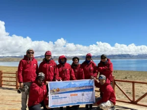 A “Kailash Mansarovar Yatra 2026” banner is displayed at Lake Manasarovar, set against blue waters, snow-capped Himalayan peaks, and dramatic clouds.