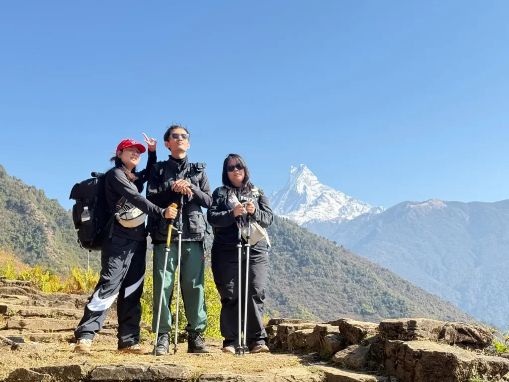Three hikers are standing on a hill, with a clear, panoramic view of the Annapurna South Himalaya behind them.