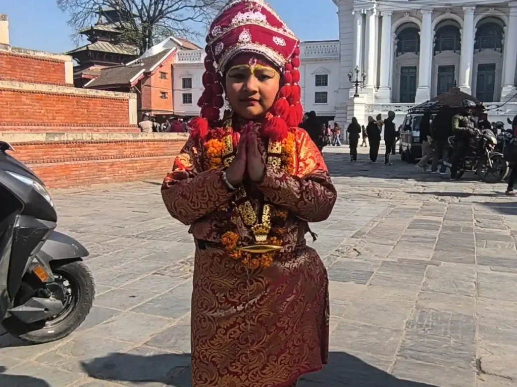 Child dressed as Kumari, the Living Goddess of Nepal, standing with Namaste gesture at Kathmandu Durbar Square near the historic palace building.
