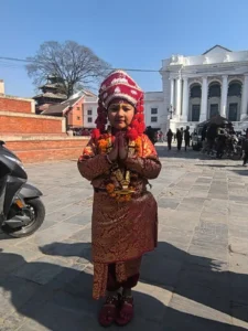 Child dressed as Kumari, the Living Goddess of Nepal, standing with Namaste gesture at Kathmandu Durbar Square near the historic palace building.