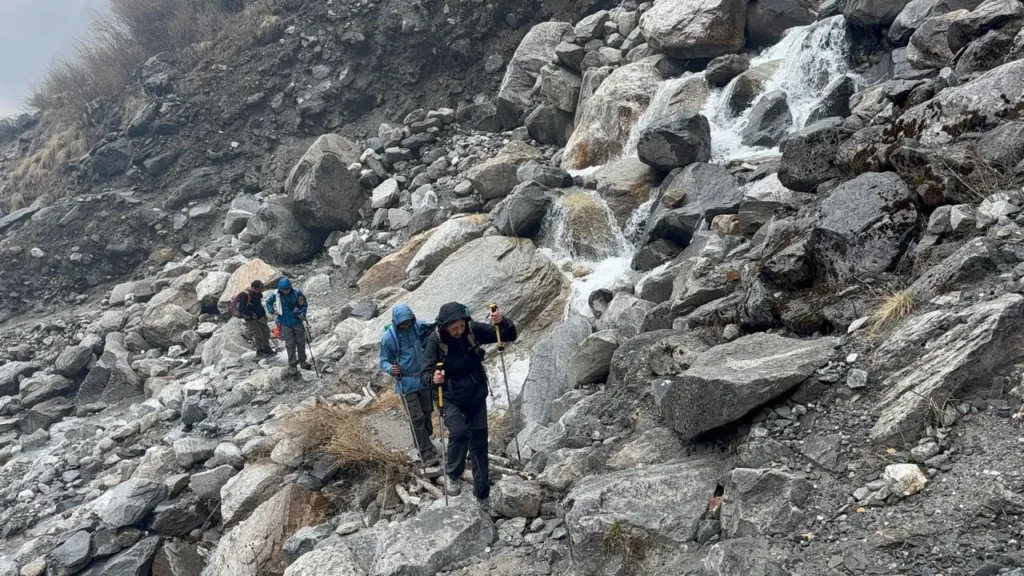 Complete Nepal Trekking Packing Guide: What to Pack & Why 6 Nepal Trekking Packing Guide: Trekkers hiking along a rocky mountain trail beside a small waterfall in a rugged Himalayan landscape